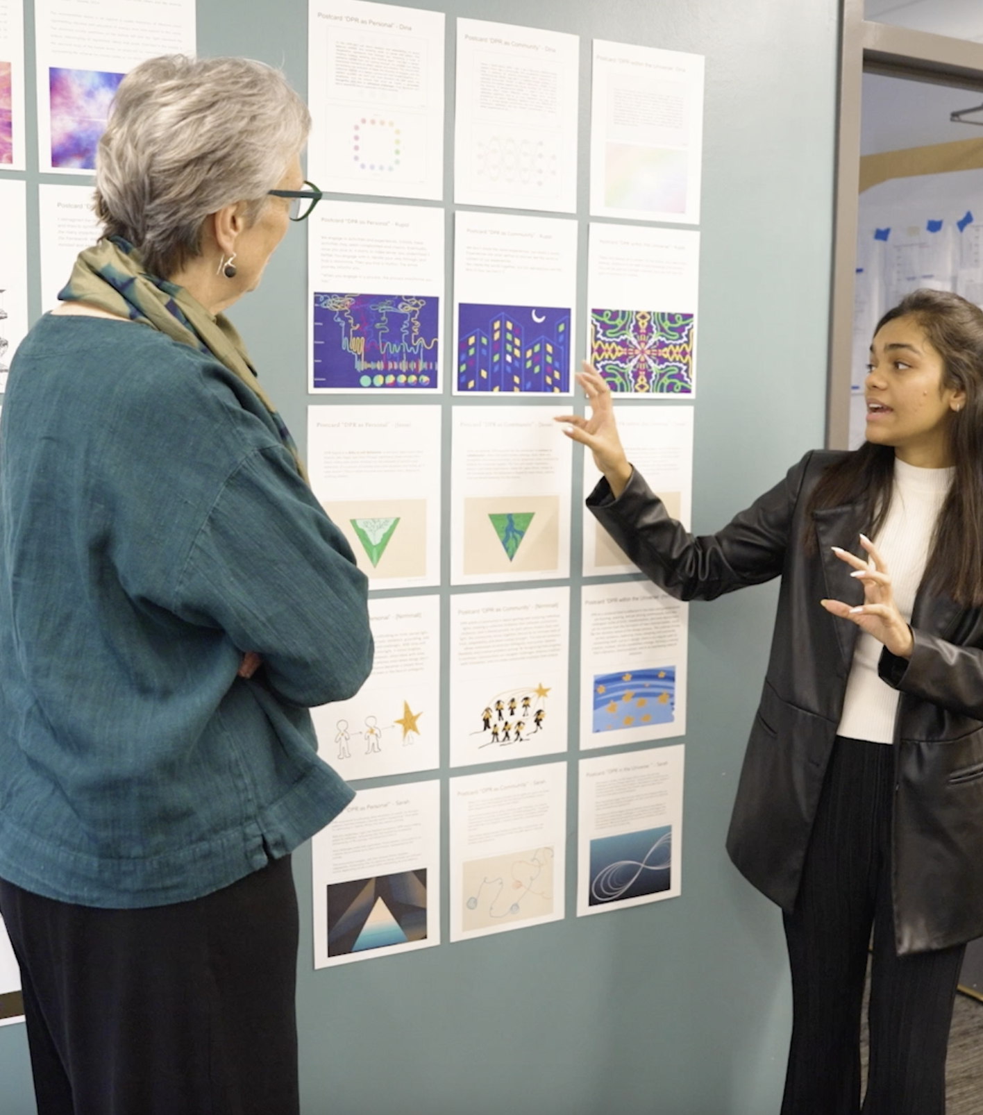 A student speaks with a HCDE professor while pointing to projects hanging on a wall