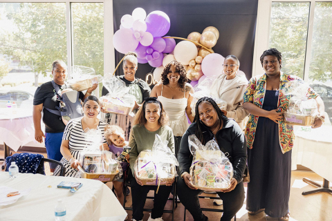 Leslie Coney with a group of attendees at the inaugural baby shower. Attendees hold gift baskets in front of a background of purple and gold balloons