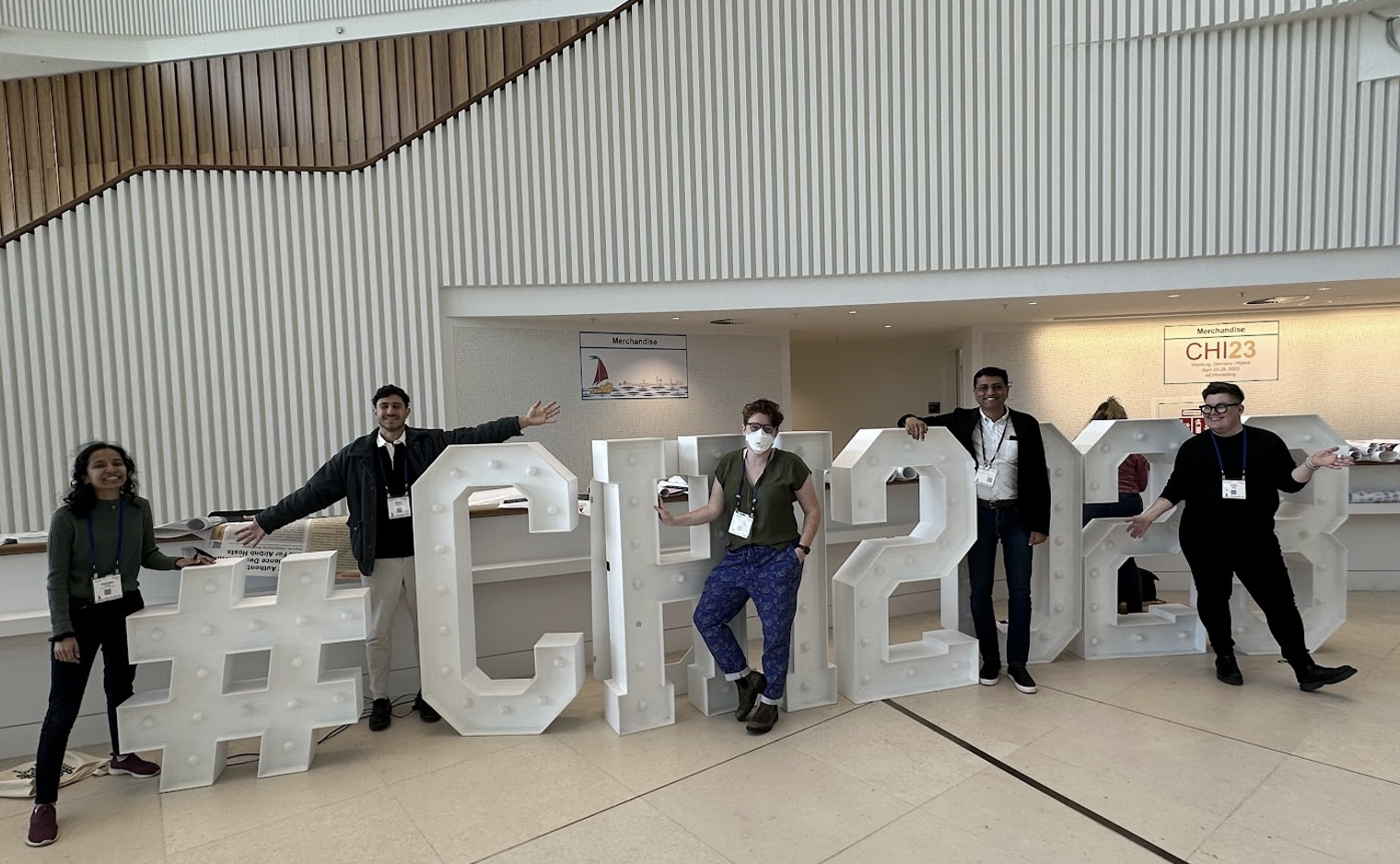 A group of 5 people stand intermixed between a set of large letters (each about 5 feet tall) that read #CHI2023, smiling and with fun poses. 
