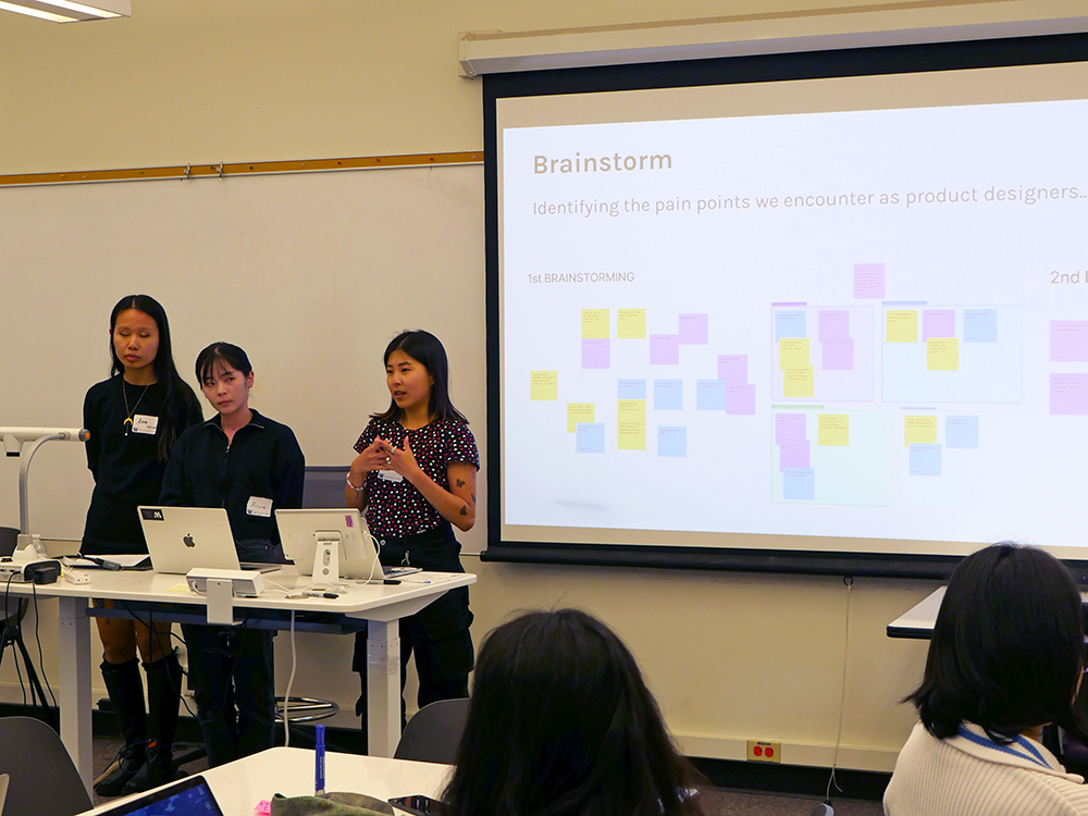 Team of three students present their prototype, standing in front of a slide that says Identifying the pain points we encounter as product designers