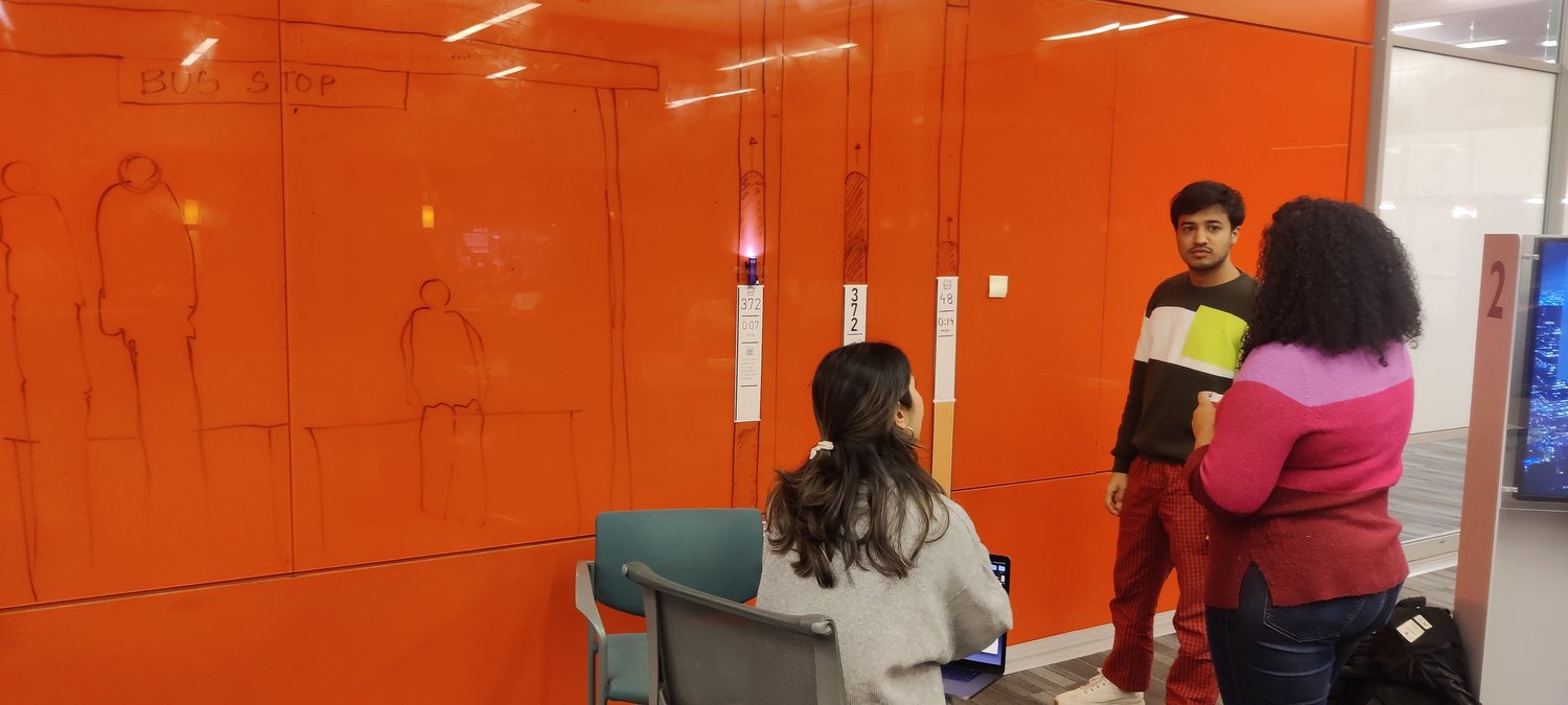 Three of students on the team in front of an orange whiteboard with sketches and paper prototypes of the Bus Beacon