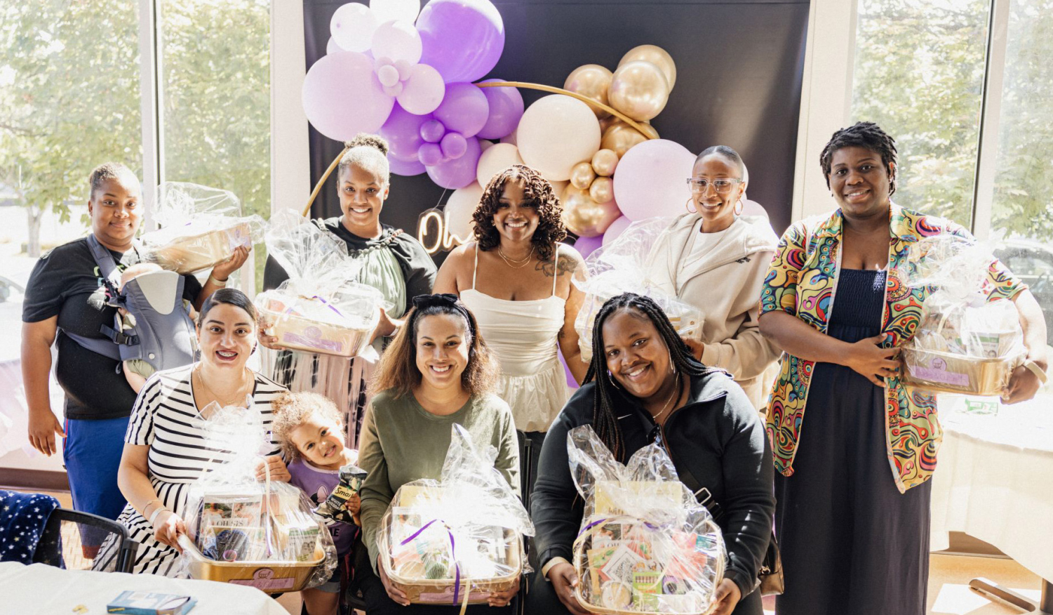 Leslie Coney with a group of attendees at the inaugural baby shower. Attendees hold gift baskets in front of a background of purple and gold balloons