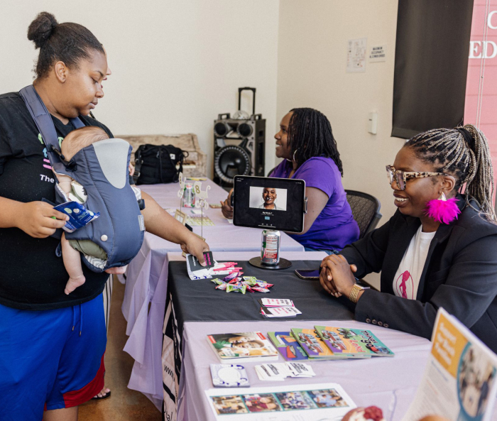 A person holding a baby on their chest stands across from someone sitting at a table with informational pamphlets