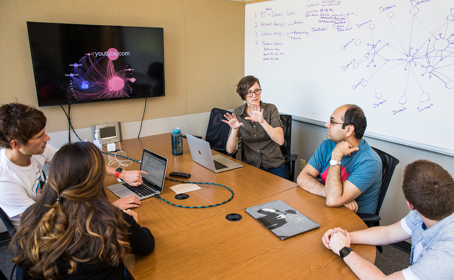 Kate Starbird sits around a table with four students. A graphic depicting information flows online is displayed on a TV and sketches are on the whiteboard behind them.