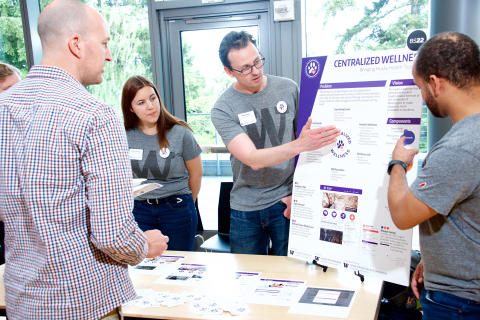 Three students with UW t-shirts behind a table pointing at their capstone project poster to a visitor